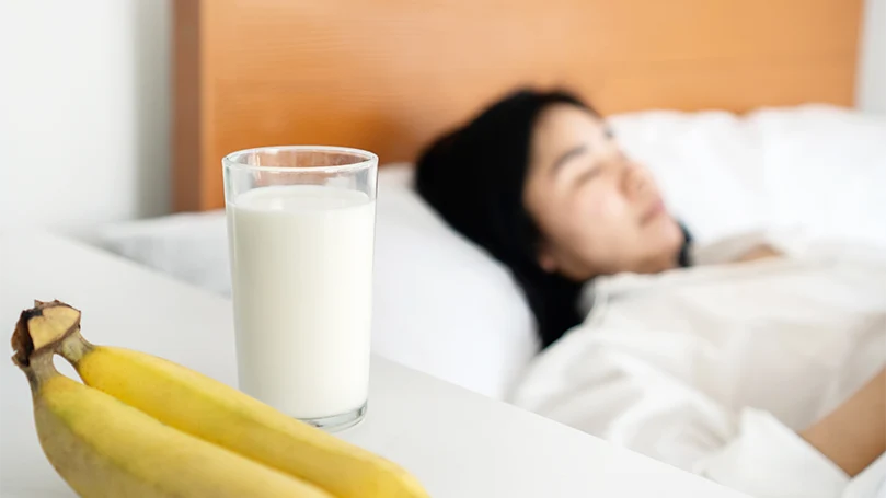 Uma imagem de uma mulher a dormir junto a uma mesa de cabeceira com leite e bananas.