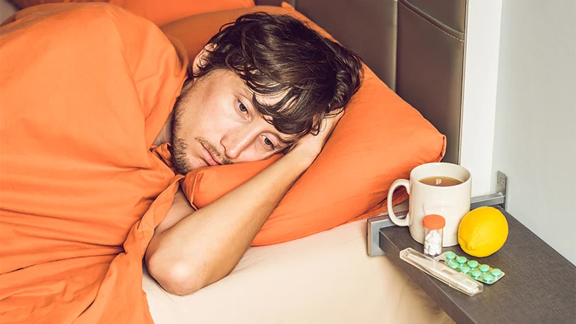 A man laying down in a bed sick and covered with an orange blanket.