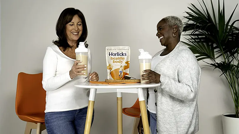 Two women holding glasses of horlicks and standing next to a table