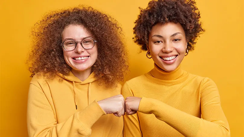 An image of two women in orange sweaters fist-bumping and supporting each other